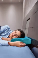 A woman peacefully sleeps on a bed, resting her head on a teal pillow. She wears a light blue sweater, conveying comfort in a softly lit room.