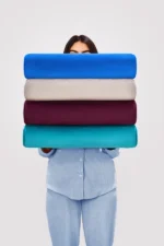 A person, mostly obscured, holding a stack of four differently colored neck pillows—blue, beige, maroon, and teal—wearing light blue button-up pajamas.