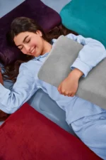 A woman in light blue pajamas smiles peacefully while hugging a gray neck pillow. She is surrounded by cozy pillows in rich red, purple, and teal tones.