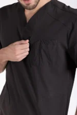 A person in a black V-neck scrub top lightly pulls on a small zipper near the chest pocket. The focus is on the zipper's details and the fabric texture.