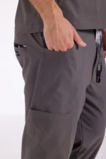 A hand rests in the pocket of gray scrub pants, featuring a pen pocket and side tag, conveying a sense of professionalism and utility.