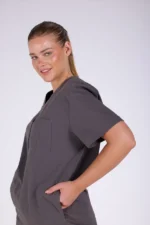 A person in gray scrubs smiles confidently, with hands in pockets. Hair is tied back, conveying a professional and relaxed demeanor. Plain background.