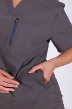 Person wearing a dark gray scrub top with a chest pocket and short sleeves. One hand is in the pocket, and the other adjusts the fabric, conveying practicality.